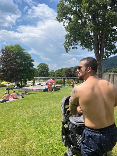 A man with tattoos on his left arm and shoulder pushes a stroller on a sunny day at a park in Oberammergau, Germany. He is wearing dark blue swim trunks with a white anchor pattern and sunglasses. In the background, people are sunbathing and swimming in a pool. Lush green grass and trees surround the area, with a cloudy blue sky overhead.