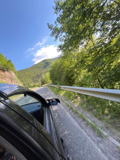 This is a photograph taken from inside a moving car on a winding road in the mountainous terrain near Taran, Bulgaria. The car's dark-colored side and open door are prominently featured in the foreground on the left. The road itself is paved and curves to the right, bordered by a metal guardrail on the right side. Lush green trees and vegetation cover the steep slopes of the mountains that flank the road. The sky is blue with scattered white clouds, and sunlight illuminates the scene. The image conveys a sense of travel through a scenic, natural environment.