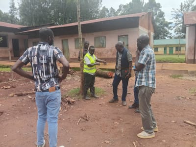 The image shows a group of five men standing in a dirt yard outside a single-story building in Idakho South ward, Kenya. The building has a rusty tin roof and faded, light-colored walls with green window shutters and doors. A sign partially visible on a second building in the background reads "UNTY GOVERNMENT OF KAKAME". One man, wearing a yellow safety vest, is measuring a length of lumber, while another man in a dark vest with a printed shirt looks on. A third man is wearing a plaid shirt. The man in the forefront is wearing a patterned shirt, blue jeans, and is standing with his arms on his hips. There is a cut log on the ground. The ground is bare earth with scattered debris. Trees are visible in the background under a cloudy sky.