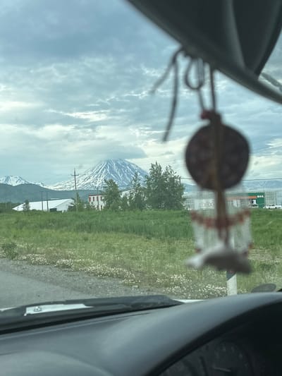 The image is taken from inside a vehicle, looking through the windshield. In the foreground, the dashboard and a portion of the steering wheel are visible. Hanging from the rearview mirror or an attachment point above it is a decorative ornament, possibly a dreamcatcher, which is out of focus.
The midground shows a landscape with green grass and a gravel road or shoulder. There are some small white flowers scattered in the grass. In the distance, there are several buildings, including a white-roofed structure, a red and white building, and larger commercial buildings with green roofs and signage. Power lines and poles are also visible.
The background features a majestic mountain range. The prominent peak in the center is snow-capped and appears to be a volcano, with a plume of smoke or steam rising from its summit. Other mountains, also partially covered in snow, are visible to the left. The sky is overcast with gray clouds.
The location is identified as Ketkino, Russia. The scene depicts a view of a natural landscape with human settlements and infrastructure, set against a backdrop of volcanic mountains under a cloudy sky.