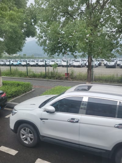 This image captures a large outdoor parking lot filled with numerous white cars, predominantly SUVs, neatly arranged in rows. The cars are positioned behind a metal fence, with lush green trees partially obscuring the view. In the foreground, a white SUV is prominently featured, its side visible from the front fender to the rear door. A smaller dark-colored car is parked to the left of the white SUV. The environment appears to be a car dealership or a storage lot, possibly located in Yufengshan, China, given the presence of a mountainous landscape in the background. The sky is overcast, suggesting a cloudy day. There are no people visible in the image.
