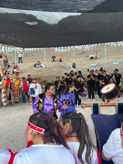 A vibrant outdoor procession features a marching band in black, a couple in ornate purple attire, and dancers in colorful traditional outfits. Spectators line the sides of the street, some watching from under a makeshift tent. Bunting hangs overhead, adding to the festive atmosphere.