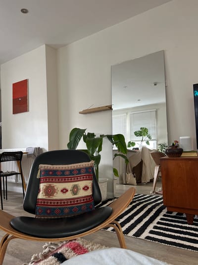 This image depicts an interior view of a domestic living space located in St Albans, United Kingdom. The foreground is dominated by a mid-century modern lounge chair featuring a black leather seat on a light wooden frame, adorned with a rectangular cushion exhibiting a geometric pattern in red, blue, beige, and white. Beneath the chair, a shaggy rug with pink and neutral tones is partially visible. The room features white walls and a light-colored wooden floor.
To the left of the lounge chair, a black slatted dining chair and a portion of a dining table are visible, with a white wall-mounted radiator below. Above this area, a rectangular abstract painting, divided into red and dark red segments with a small white circular detail, hangs on the wall.
Towards the right, a large, unframed floor-length mirror stands against a white wall. Reflected in the mirror are a window with horizontal blinds, a large green houseplant, and a section of a room beyond. A large, dark green-leafed houseplant in a woven basket pot is situated in front of the mirror. Above the plant, a narrow wooden shelf holds several incense sticks.
On the far right, a wooden sideboard supports a dark, patterned ceramic bowl, a stack of books, and a white speaker. A portion of a television screen displaying blue and green light is visible on the extreme right edge. A black and white striped textured rug is laid on the wooden floor in the midground, extending towards the reflected area. No individuals or active interactions are present within the scene.