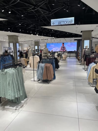 Here is a forensic description of the image:
**Location:** The image was taken in a clothing store in the city of Саларьево, Russia.
**Setting:** The scene is an aisle of the store, which is filled with racks of clothing. The floor is white tile. Above, the ceiling is a network of black architectural elements and spotlights. There is a sign, written in Russian, with an arrow that points left, saying "Детский" (which translates to "Children's"). In the background there is a large display screen, showing an image of a man against a cloudy blue and white background.
**Objects:**
* Racks of clothing in a variety of styles and colors
* A sign with an arrow
* White tile floor
* Large display screen
* Black structural supports
**Inferences:**
Based on the clothing and the sign, it is likely that this is the children's section of the store.