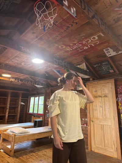 A young woman stands in a rustic room, possibly a cabin or bunkhouse, with wooden paneling on the walls and ceiling. The ceiling is covered in graffiti, including names, dates, and drawings, and features a basketball hoop. A skylight illuminates the room. Two simple wooden beds with mattresses and pillows are visible. A wooden door covered in more graffiti, including "Yahi Guy", stands to the right. The woman, wearing a yellow oversized t-shirt and brown pants, is touching her hair and looking to her right, appearing to be mid-motion. The scene is set in Fryeburg, United States of America.