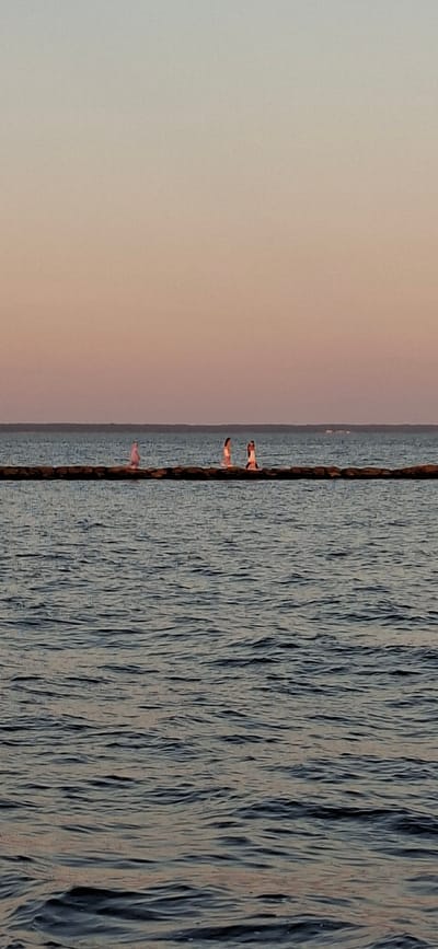 This image depicts a serene waterscape at dusk, with a clear horizon separating the calm sea from a pale pink and orange sky. In the foreground, the water exhibits gentle ripples, reflecting the soft colors of the sunset. A long, dark structure, possibly a breakwater or pier, stretches horizontally across the middle ground. On this structure, three individuals are visible, standing some distance apart. The two figures on the right appear to be women dressed in light-colored clothing, possibly swimwear, with one of them raising her arm. The figure on the left is more indistinct, also in light clothing. The background shows a faint, dark line that could be the distant shoreline of Pärnu linn, Estonia. The overall atmosphere is tranquil and atmospheric, capturing the peaceful end of a day by the sea.