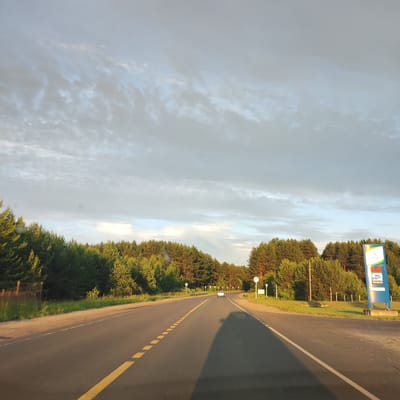 This media file captures a view from a vehicle traveling on a paved road lined with trees, likely in Kineshma, Russia. The scene is set in a rural or semi-rural area, with dense coniferous and deciduous forests bordering the road on both sides. The road itself is asphalt and marked with yellow dashed lines, indicating a divided lane. In the distance, a white car is visible, suggesting ongoing traffic.
The sky is a prominent feature, filled with a dramatic display of clouds, with patches of blue sky peeking through. The lighting suggests either early morning or late afternoon, with the sun casting long shadows across the road and illuminating the trees with a warm glow. The weather appears to be partly cloudy, creating a dynamic and visually interesting atmosphere.
On the right side of the road, a blue sign with a logo and some text is visible. While the text is not entirely clear, parts of it appear to be in Cyrillic script. A digital display on the sign shows a time, "23:35:18," and what appears to be a price, "969.00". There are also smaller white signs along the roadside, possibly indicating speed limits or other traffic information. The presence of a fence near the left edge of the frame suggests the proximity of property or perhaps agricultural land. The overall impression is one of a journey through a scenic, natural landscape under a dynamic sky.