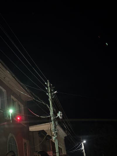 This image depicts a nighttime urban street scene in Bazan, Bulgaria. The environment is dark, illuminated by artificial light sources.

A prominent wooden utility pole stands centrally, heavily laden with numerous grey electrical and communication wires stretching in various directions across the frame. Multiple insulators and fixtures are attached to the pole, along with a small, white rectangular object secured mid-height.

To the left, a multi-story building with a reddish-brown facade is partially visible. It features at least one window with an arched top and a white cornice below the roofline. A bright green exterior light is mounted on this building, and directly below it, a distinct red light source is also present, possibly part of a traffic signal or architectural lighting.

To the far right, a modern street light with a bright white-green glow illuminates the lower right portion of the scene. In the upper right quadrant of the dark sky, two faint, small light sources are visible: one red and one green.

At the extreme bottom edge of the frame, indistinct dark shapes consistent with the tops of human heads are discernible, indicating the presence of individuals, though no specific features, actions, or interactions are visible.