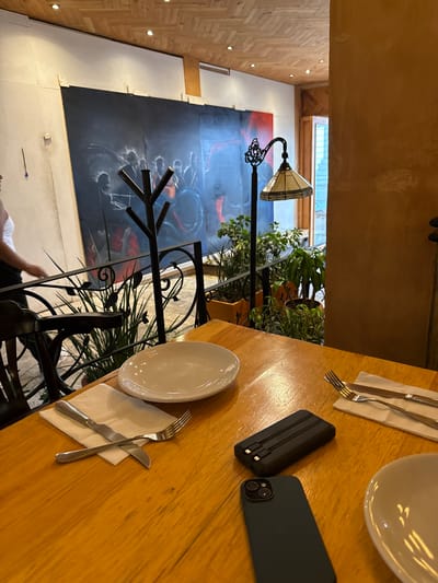 This is an indoor shot of a dining table at a restaurant in San Cristóbal, Mexico. The table is wooden and set with plates, silverware, and napkins. A black smartphone and a black portable charger are placed on the table. A decorative wrought iron railing and plants are partially visible near the table. In the background, there's a lamp and a large painting on the wall. A glimpse of a person in a white shirt can be seen near the bottom of the frame. The overall environment appears to be a restaurant setting with an artistic and natural ambiance.