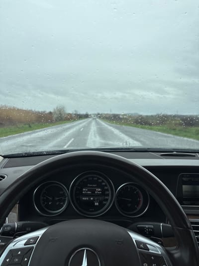 The image depicts the interior of a Mercedes-Benz car on a wet road, presumably in transit. Raindrops are visible on the windshield, obscuring the view of a two-lane road stretching into the distance under an overcast sky. The car's dashboard is in view, including the speedometer which reads 57 km/h. The temperature is 10.5°C, and the car is in drive ("D"). The navigation system is showing "Navi" in the top left corner. Based on the OCR, the image was taken at 12:49 PM. The city is listed as Laka, Bulgaria. No people are visible.