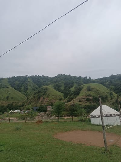 The image depicts a natural landscape with rolling green hills under a cloudy sky. In the foreground, there is a grassy area with a dirt patch, possibly a volleyball court, and a white yurt with decorative patterns. There are also some wooden poles and a net. In the middle ground, a cluster of buildings and more yurts are visible, along with some trees and rocks. The background is dominated by lush green hills covered in vegetation. The scene appears to be set in a rural or nomadic settlement.