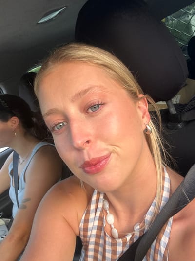 The image displays two females inside a vehicle. The primary subject, a fair-skinned female with blonde hair, light blue eyes, and light makeup including blush and lip gloss, occupies the foreground. She wears a brown and white gingham check halter-neck top, a necklace with several large, light-colored irregular beads, and a silver-toned hoop earring on her visible right ear. A dark seatbelt diagonally crosses her torso. She is looking directly towards the camera with a slight smile. To her left and slightly behind, a second female with dark hair, styled with a black wavy headband, is partially visible from her left profile. She wears a light-colored top and a delicate necklace. The car's interior features a dark headrest behind the foreground subject and a light-colored ceiling with an integrated light fixture. Through a rear window, faint greenery is visible in the background, suggesting the vehicle is outdoors. The scene is illuminated by strong natural light. The specific location within Thirroul, Australia, is not identifiable from the visual content of the image.