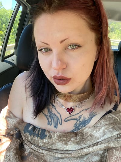A woman with green eyes and two-toned hair (red on top, black underneath) is looking at the camera. She has tattoos on her chest and is wearing two necklaces. She is inside a car; the window shows green trees and a bright sky. The location, Kruče-Kërruçi, Montenegro, is not directly visible.