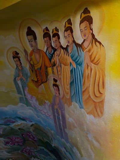 This is a detail of a colorful mural, likely religious in theme, depicting several figures with halos and serene expressions. The figures are depicted in a profile or semi-profile view, with some holding their hands in a prayer-like gesture. They are adorned in flowing robes of vibrant colors such as orange, yellow, blue, and purple. In the foreground, a lush scene of lotus flowers and large lily pads unfolds, rendered in shades of pink, purple, and green, set against a backdrop of swirling clouds or mist. The overall impression is one of spirituality and tranquility. This mural is located on the exterior wall of the Linh Phuoc Pagoda in the city of Nam Ban, Vietnam.