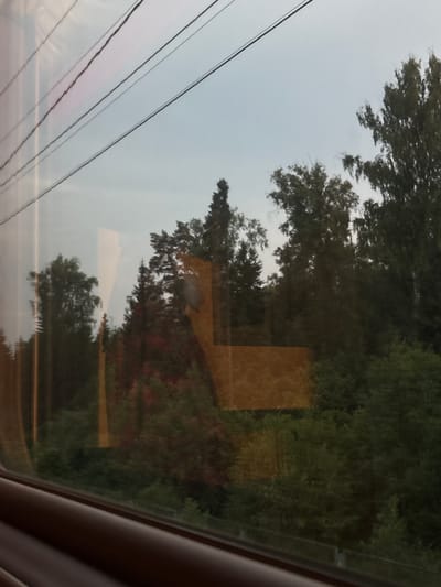 The image captures a view from a train window, looking out at a wooded area and a light sky. Three overhead wires are visible, running diagonally across the upper portion of the frame. The reflection on the window shows trees and a pale sky. A rectangular, orange-tinted reflection overlays a section of the trees. The lower part of the image shows the dark brown interior of the train, including the window frame and a portion of the seat. The scene appears to be on a clear day. There is no indication of the specific location in Povarovo, Russia.