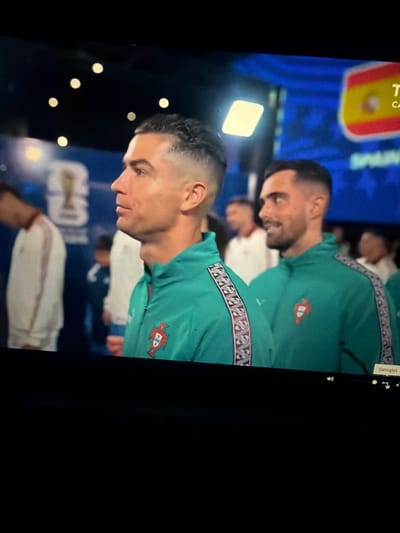 The image captures a close-up of two men wearing matching teal athletic jackets with a patterned trim. The man on the left, in the foreground, is identified as Cristiano Ronaldo due to his distinctive facial features and haircut. He is looking to his left, with a serious expression. He is wearing a teal jacket with a Portuguese crest on the left chest. The man to his right, partially visible, is also wearing a similar jacket and looking towards his left with a slight smile. The background is blurred and appears to be an indoor setting with blue and yellow banners, one of which displays the Spanish flag. There are also out-of-focus figures in the background. The lighting suggests an artificial environment, possibly a stadium or an event venue. The image is a still frame from a video, as indicated by the presence of video playback controls at the bottom right, including a "Genişlet" button, which is Turkish for "Expand." The image does not contain any information indicating a location in Nikopol, Bulgaria.