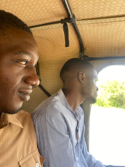 In Yali, Nigeria, two men are seen from the chest up, seated inside a vehicle, likely an auto-rickshaw, given the context of the location. The man in the foreground, positioned on the left, has dark skin and is looking forward, with his head turned slightly towards the viewer. He is wearing a tan shirt with visible stitching and a button. The second man, on the right, also has dark skin and is looking out of the vehicle's window. He is wearing a striped shirt. The interior of the vehicle features a checkered ceiling. The scene is illuminated by natural light, suggesting it is daytime.