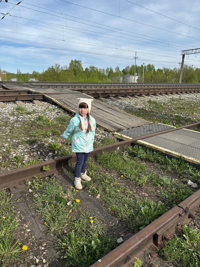 A young girl stands on a railway track in Кечёво, Russia. She is wearing a light blue jacket with a unicorn pattern, dark blue leggings with white polka dots, and light beige shoes. A white hat with animal ears is on her head. The railway track is overgrown with grass and small yellow flowers. A wooden walkway with a grey, textured section runs across the tracks. Railway lines and power lines are visible in the background. The sky is partly cloudy.
