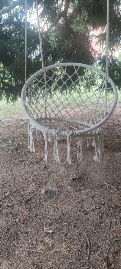 This media file shows a white macrame hammock chair hanging from the branches of a large tree. The chair is oval-shaped with a diamond-patterned weave and fringe tassels hanging from its bottom edge. Two thick ropes suspend the chair from the tree. The surrounding setting is outdoors, with dry, brown pine needles and small branches covering the ground beneath the chair. Lush green foliage from the tree forms a canopy overhead, and in the background, through the branches, a building with a reddish-pink facade and white window frames is partially visible. The lighting suggests it is daytime, possibly overcast or late afternoon, as there are no strong shadows. The scene appears to be a tranquil, natural environment, likely a backyard or park in Liebenburg, Germany, given the context. There is no text visible in the image.