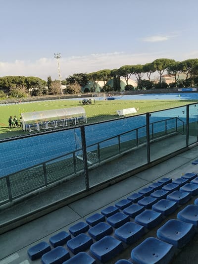 The image displays an outdoor multi-sport stadium, identified as the Stadio Comunale Italo Nicoletti in Riccione, Italy, captured from an elevated spectator seating area on a clear day. The foreground shows multiple rows of empty, blue plastic stadium seats, positioned above a glass safety barrier with metal railings that separates the seating from the track level.
In the midground, a bright blue synthetic running track encircles a neatly maintained green grass sports field. On the grass, near the center-left, a cluster of approximately 10-15 individuals, some attired in dark athletic wear, are gathered in a close huddle. To their left, a smaller group of 3-4 individuals in athletic gear stands near yellow cone markers. A clear, semi-transparent player dugout structure is situated alongside the track on the left. White lines delineate sections of the grass field, and a goalpost is visible at the far end. A partially obscured white banner is located along the far edge of the blue track.
The background is dominated by a distinct line of tall, dense green-canopied trees, likely umbrella pines, forming a natural boundary around the stadium. Beyond the trees, sections of buildings and a large white domed structure are visible. A prominent, tall floodlight pole equipped with multiple lamps stands on the left side, extending into a light blue sky with scattered white clouds. The scene is illuminated by bright natural daylight.