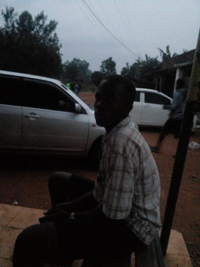 The scene is an outdoor environment in East Ugenya ward, Kenya, captured under low light conditions, indicative of dusk or heavy overcast. In the immediate foreground, a dark-skinned adult male is seated on a light-colored concrete surface, facing towards the right of the frame but looking slightly towards the camera's position. He is wearing a light-toned plaid short-sleeved shirt and dark trousers; a watch is visible on his left wrist. Behind him, on an unpaved, reddish-brown dirt road, a silver five-door hatchback or small SUV is present, with its driver's side visible. Further into the mid-ground, a blurred adult male figure is in motion, walking from right to left, dressed in a light-colored top and dark trousers. A second white vehicle is partially visible behind this moving figure. The background features dense green foliage, including trees, and a basic structure with a brown roof is discernible on the far right. Overhead power lines stretch across the dull, grey sky. A dark, vertical pole stands in the extreme right foreground.
