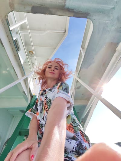 The image shows a woman with reddish-blonde hair taking a selfie outdoors. She is wearing a colorful, patterned shirt. Her left arm is extended towards the camera. The background appears to be the deck of a ferry or ship, with white railings and structures visible. The sky is blue and sunny, with some lens flare from the sun. The scene is not located in the city of Nova, Russia, as there is no information within the image to suggest this location.