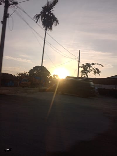 The image captures a sunset scene in Bintang Meriah, Indonesia. A tall palm tree stands silhouetted against the bright sky, with power lines crisscrossing overhead. In the midground, a dump truck is parked near some buildings, and other vehicles are visible. The sun is low on the horizon, casting a warm glow and lens flares across the scene. The foreground is a dark road, suggesting the image was taken from a moving vehicle. The word "umi" is visible in the bottom left corner, possibly a watermark or signature.