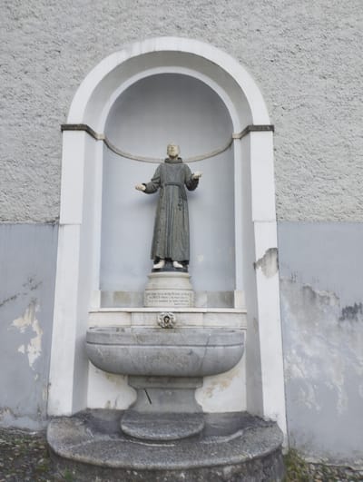 A statue of a monk in a dark robe stands in a niche on the exterior wall of a building in Orselina, Switzerland. The statue is positioned above a stone fountain. The wall behind the niche is textured and painted light gray. The niche itself is arched and framed by white trim. Below the niche, a stone fountain bowl is mounted on a pedestal, with steps leading up to it. The overall scene appears to be outdoors, with some grass and dirt visible at the base of the structure.