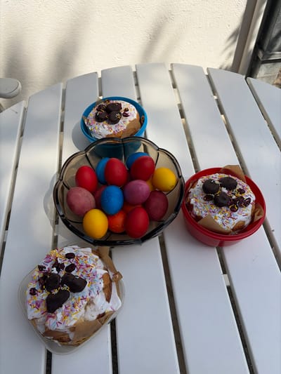 The image shows a white, slatted table with Easter treats arranged on it. There are three round cakes decorated with white icing, sprinkles, and dark chocolate pieces. One cake is in a red container, one in a blue container, and one in a clear square container. In the center of the table, a dark, bowl-shaped container holds a collection of colorful Easter eggs in shades of red, blue, yellow, purple, and orange. The setting appears to be outdoors, possibly on a patio. The location is in Topola Municipality, Serbia.