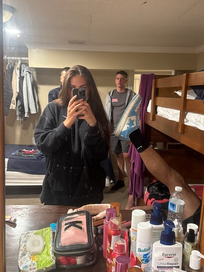 This is a mirror selfie taken in a dorm room, likely at a motel or hotel in Ocean City, Maryland. In the foreground, a young woman is taking the photo with her phone, wearing a black hoodie. Her long brown hair obscures part of her face. To her right, in the reflection, a young man in a grey t-shirt and shorts stands near a bunk bed. Another young man is partially visible behind the woman. A third person's legs are visible in the reflection, with one leg raised and wearing a blue and white sneaker. On the table in front of the mirror are various toiletries, including baby wipes, a makeup bag, and several bottles of lotion and hair spray. The room appears to be a shared living space with bunk beds and a closet filled with clothes.