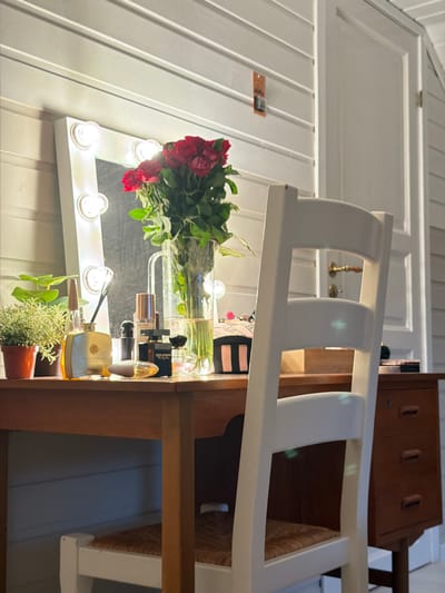 The image shows a vanity table with a lighted mirror, a vase of red roses, several perfume bottles, and a small potted plant. A white chair with a woven seat is pulled up to the table. To the right of the vanity is a brown wooden dresser with three drawers. The background features a white paneled wall and a white door with a brass doorknob. The scene appears to be an indoor setting, possibly a bedroom or dressing area, located in Kleppe, Norway. There are no people visible in the image. The lighting is warm, likely from the vanity lights and possibly overhead room lighting. The overall impression is one of a well-appointed and tidy personal space.