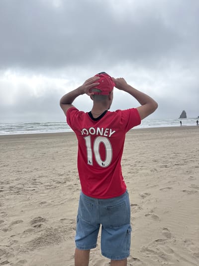 A man stands on a sandy beach, facing away from the camera. He wears a red soccer jersey with the name "ROONEY" and the number "10" on the back, along with denim shorts and a red baseball cap. He appears to be holding his head with his hands. The backdrop consists of a cloudy sky and ocean waves crashing on the shore. A few distant figures are visible further down the beach.