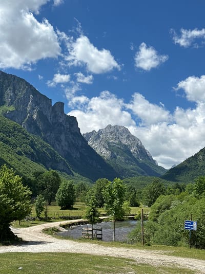 This is a wide shot of a rural landscape in Vusanje, Montenegro, under a bright blue sky with scattered white clouds. In the foreground, a dirt road curves from left to right, bordering a grassy area. A shallow river runs across the middle ground, with a small wooden structure resembling a footbridge or dam. Beyond the river, more dirt paths and grassy fields are visible, dotted with trees. The background is dominated by lush green mountains, with rugged, rocky peaks visible in the distance. A small blue sign with white lettering is visible on the right side of the image. The overall scene is tranquil and natural, with no people visible.