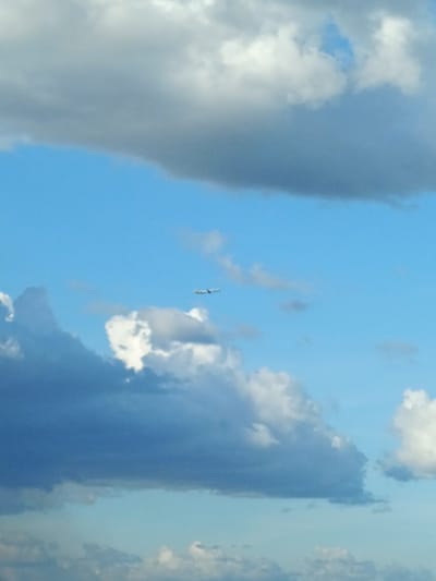 The image shows a clear blue sky with white and gray clouds. A single airplane is visible in the upper middle of the frame, flying from left to right.  The sky is bright and the clouds appear fluffy and spread out. The location is Parañaque City, Philippines, at coordinates (latitude: 14.474, longitude: 121.008).