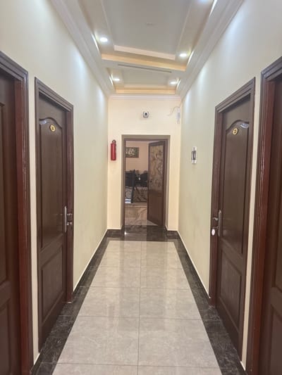 This image depicts an empty interior hallway located in Aragats, Armenia. The hallway features cream-colored walls and a reflective tiled floor composed of light grey squares bordered by dark grey tiles. The ceiling is multi-tiered with recessed sections and flush-mounted circular LED spotlights.

Dark wooden doors with silver handles line both sides of the corridor. On the left, two full doors are visible, with the closer one displaying a gold-colored oval plaque bearing the number "24." On the right, one full door is visible, also with a gold-colored oval plaque, this one marked "23."

At the far end of the hallway, an open doorway reveals a glimpse into an adjacent room, where dark furniture, possibly tables or desks, and a framed picture on the far wall are discernible. Above this doorway, a black security camera is mounted on the wall. A red fire extinguisher is mounted on the left wall of the hallway, approximately halfway down. On the right wall, directly opposite the fire extinguisher, a white rectangular wall-mounted device is present. No individuals are visible within the scene.