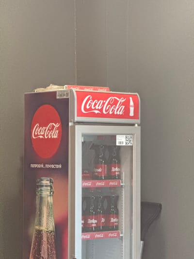 A refrigerated Coca-Cola vending machine stands indoors, likely in a shop or public space. The machine features Coca-Cola branding prominently, with the company's logo displayed in both English and Russian. The interior is filled with bottles of Coca-Cola, each featuring the Russian word "Добрый" (Dobryy), which translates to "Good" or "Kind." A QR code is visible on the machine's frame. A promotional image below the cooler section shows a bottle of Coca-Cola and the phrase "ПОПРОБУЙ... ПОЧУВСТВУЙ" (Poprobuy... Pochuvstvuy), meaning "Try... Feel". Based on the information available, the scene is most likely in Novo-Nikol'skoe, Russia.