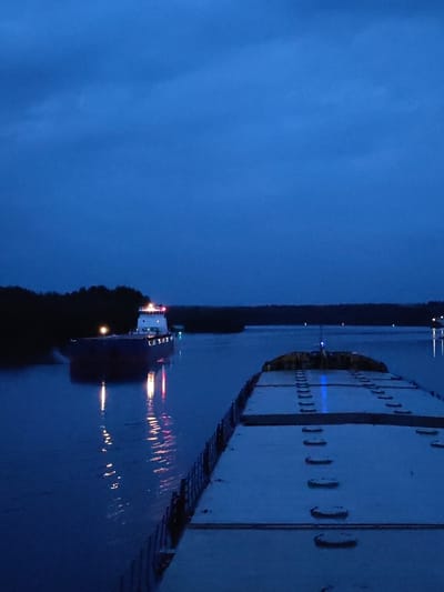 The image depicts a dusky scene on a body of water, likely a river or canal, near the city of Yalosar', Russia. The sky is a deep, muted blue, suggesting either dawn or dusk. In the foreground, the stern of a large barge is visible, its deck covered in metal gratings and hatches. The barge is moving forward, indicated by the wake it leaves on the water.
Further ahead, on the left side of the frame, another vessel, possibly a tugboat or a small cargo ship, is approaching. This vessel is illuminated with red and white lights, and its reflection shimmers on the dark water. The water itself appears calm, with gentle ripples. Along the distant bank, a dense line of dark trees forms a silhouette against the twilight sky. The overall atmosphere is quiet and serene, conveying a sense of the vastness of the waterway and the journey of the vessels.