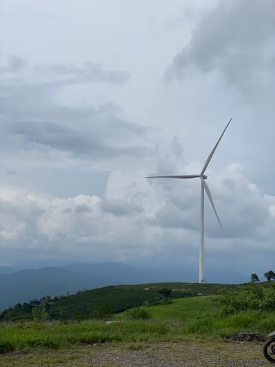 The image depicts a wind turbine standing tall on a grassy hillside in Trường Thọ, Vietnam. The sky is overcast with dramatic, gray clouds. In the foreground, a gravel path leads towards a lush green landscape dotted with trees and rolling hills. Several layers of distant mountains recede into the hazy horizon. A portion of a motorcycle is visible in the lower right corner of the frame.
