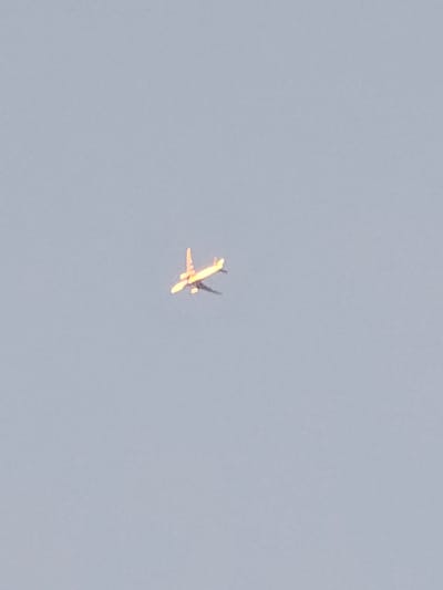 An airplane is pictured in the sky above Mäntsälä, Finland. The airplane is illuminated by the sun, giving it a golden hue. The sky is a pale blue, with no clouds visible. The airplane is captured from below, with its wings and fuselage clearly visible. The image is taken from a distance, and the airplane appears small against the vastness of the sky.