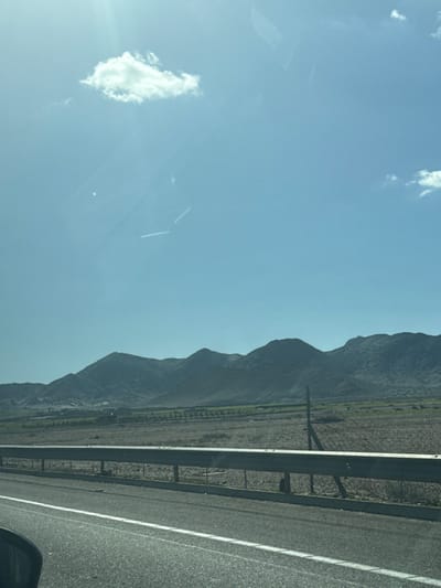 The image presents a wide landscape view from a moving vehicle on a paved road in Jbilate الجبيلات, Morocco. The foreground features the dark interior of a vehicle visible in the bottom-left corner, alongside an asphalt road marked with a solid white line on the left and a dashed white line to its right. A silver metal guardrail, supported by dark posts, runs horizontally across the lower mid-ground, separating the road from the undeveloped terrain beyond.
The mid-ground consists of expansive, mostly barren, light brown ground with patches of low, green vegetation. Rows of what appear to be cultivated plants or small trees are discernible in the left-center mid-ground. A tall, slender dark pole stands upright in the field behind the guardrail, slightly right of the frame's center.
In the background, a range of undulating, dark grey hills or mountains dominates the horizon under a bright, clear light blue sky. Sparse lighter-toned areas are visible on the mountain slopes. The sky contains a single distinct white cumulus cloud in the upper-left quadrant and less prominent cloud fragments near the upper-right. Sun flare is present in the upper-left, and faint linear reflections, resembling contrails or internal vehicle elements, are visible across the sky portion of the image. No individuals or direct human interactions are depicted.