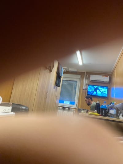 The image shows a man sitting at a table in what appears to be a room with wooden walls. He is wearing a green t-shirt and is looking towards a television screen mounted on the wall, which is displaying a video game. On the table in front of him are several water bottles, a banana, and a small container. To the left, there is a white electronic device and a black suitcase. There are also clothes hanging on a rack on the wall. The room is lit by a fluorescent light fixture on the ceiling. There is a window with a blue blind behind the man, and a radiator below it. The overall scene suggests a casual, possibly recreational setting.