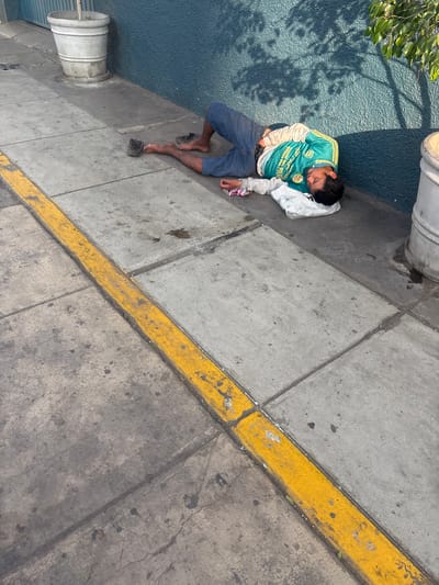 A man lies on a sidewalk in La Victoria, Peru. He is wearing a green and yellow soccer jersey, blue shorts, and a white long-sleeved shirt. He is barefoot. He is lying next to a blue wall and a metal object that might be a trash can. A yellow line runs along the sidewalk.