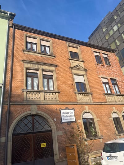 An upward-angled outdoor shot captures the facade of an old brick building under a clear blue sky. The building, constructed with light reddish-brown bricks, features multiple stories and ornate architectural details around its windows. Rectangular windows on the upper floors have decorative stone surrounds, some with external roller blinds and others with white internal curtains.

On the ground floor, a large, arched wooden entrance door with a patterned glass fanlight is visible. To the left of the door, the house number "16" is affixed to the brick wall. Above and to the right of the entrance, a white, rectangular sign with a black decorative border reads "Albert Eb. Metallgießerei Armaturenfabrik" (Albert Eb. Metal Foundry Valve Factory). A small, yellow sign on the entrance door reads "Einfahrt freihalten" (Keep entrance clear).

Adjacent to the building's entrance, a patch of bare-branched shrubs grows, and a brown outdoor utility box or mailbox is present. A white Toyota Yaris hatchback is partially visible, parked on the street to the right, displaying a German license plate "SU:XQ 826". In the upper left background, part of a modern building with a glass grid facade reflects the blue sky. The scene is brightly lit by daylight, suggesting good weather. No people are visible. The location context for the building is Schweinfurt, Germany.