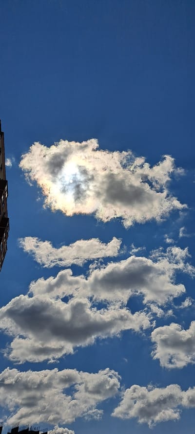 A low-angle, vertical photograph captures a bright blue sky filled with various clouds. The sun is partially obscured by a large, iridescent cloud in the upper middle portion of the frame, creating a halo effect with rainbow hues. Below this, several other fluffy white and grey clouds are scattered across the sky. In the lower left corner, the edge of a tall, dark-colored building is visible, and in the bottom edge of the frame, the dark, skeletal framework of a construction site is present. The scene appears to be an urban environment, potentially the city of Zhukovo, Russia, given the context. There are no people or animals visible in the image.