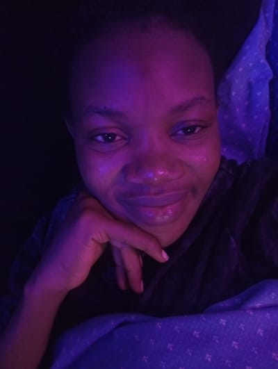 A close-up, self-portrait photograph shows a young Black female with dark hair and brown eyes, lit by a purple light. She is resting her chin on her hand, which is positioned at her jawline. Her lips are slightly parted, and she is looking directly at the camera with a gentle smile. She is wearing a dark-colored garment, possibly a hoodie. The background is dark, with a section of blue fabric with a white pattern visible to the right. The scene appears to be indoors, possibly in a bedroom, with the purple lighting creating a moody atmosphere. The image was taken in Ashipa, Nigeria.