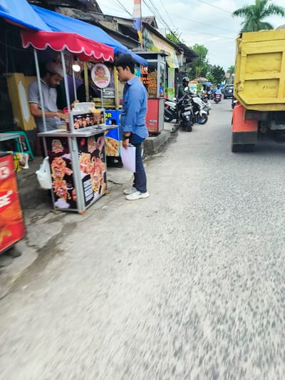 The image shows a street scene in Kecamatan Dayeuhkolot, Indonesia, located at latitude -6.963 and longitude 107.595. A man in a blue shirt and white sneakers is standing in front of a food stall, looking at a food item in a container. Another man in a light-colored shirt and black pants is standing behind the stall, looking towards the man in blue. The stall is made of metal and has a blue and red awning, displaying a picture of food. The street is paved and has a light gray color. Several motorcycles are parked on the side of the street, and a yellow truck is driving away. There are some buildings in the background, all painted with a variety of colors. The sky is cloudy.