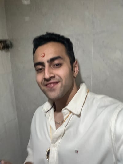 A young adult male is depicted from the chest up, positioned slightly right of center, against a plain light-grey tiled wall. He has short dark hair, dark eyebrows, and a light complexion, exhibiting a closed-mouth smile. A reddish-orange circular mark, possibly a tilak, with a darker central symbol, is visible on his forehead. A small silver-colored hoop earring is present in his right earlobe. He wears a light-colored, possibly off-white, collared shirt, unbuttoned at the top, revealing a small flag logo on the left chest area. A thin gold-colored chain is visible around his neck, and a silver-colored chain with a dark, oval-shaped pendant hangs over his shirt. The lighting is uniform. The scene takes place in Bidhannagar, India.