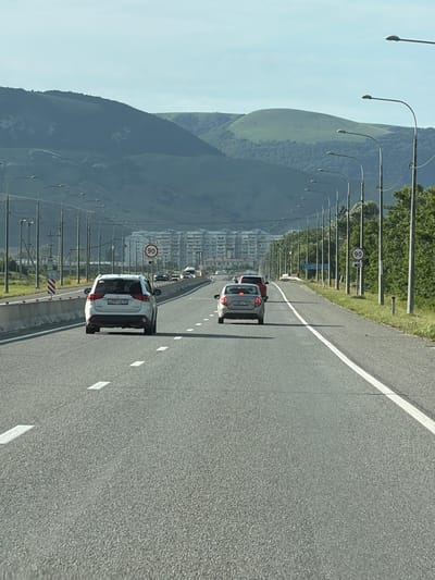 The image shows a view from a moving vehicle on a highway. The highway is paved and has white lane markings. On the left side of the highway, there is a steep, tree-covered mountain range. A row of streetlights lines the side of the road, curving upwards towards the mountain. In the distance, there are buildings that appear to be part of a city. Several cars are visible on the highway, all traveling in the same direction. One car, a white SUV, is in the foreground, and another car, a red hatchback, is behind it. There is a speed limit sign visible on the left side of the road, indicating "90". The sky is a light blue, suggesting it is daytime with clear weather. The scene appears to be set in a mountainous region with an urban area nearby. The license plate on the white SUV is visible and appears to read "A091CB 92". The location context is given as Kubina, Russia. There are no people clearly visible in the image. The overall impression is one of travel through a scenic and developed area.
