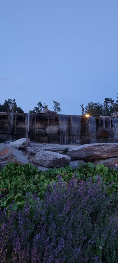 A man-made waterfall cascades over a rough stone wall, with dark green foliage and purple flowers in the foreground. The sky is a clear, pale blue. A single yellow light fixture is visible behind the waterfall on the right side. Trees are silhouetted against the sky above the waterfall.