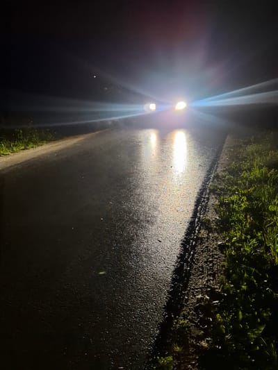 The image depicts a wet asphalt road at night, illuminated by the headlights of an approaching vehicle. The vehicle's lights create strong glare and reflections on the wet surface. The roadside on the right side of the image features grass, and on the left, it appears to be a similar grassy verge. The sky is dark and cloudy, with a faint light source, possibly a star or distant light, visible above the glare. The location is identified as Novlyanka, Russia.